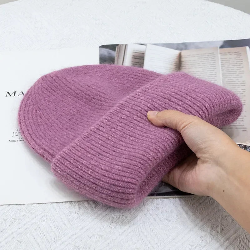 Une main tenant un Bonnet Angora Pour Maman plié et doux, violet, posé sur un magazine ouvert, placé sur une surface blanche texturée.