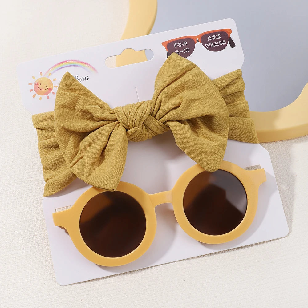 Bandeau bébé jaune avec nœud et lunettes de soleil rondes assorties, ou Set d'Accessoires Bébé | Lunettes de Cheveux pour Bébé, de BABY PREMA, conditionnées sur une carte présentoir en carton avec une illustration arc-en-ciel et soleil.