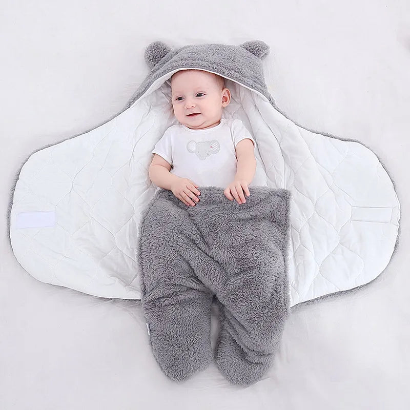 Un bébé souriant allongé sur une douce couverture en polaire Ultra Doux et Moelleux grise avec des oreilles d'ours, blotti dans une jolie gigoteuse en forme d'ours, entouré du nécessaire pour bébé. (Marque : BÉBÉ PREMA)
