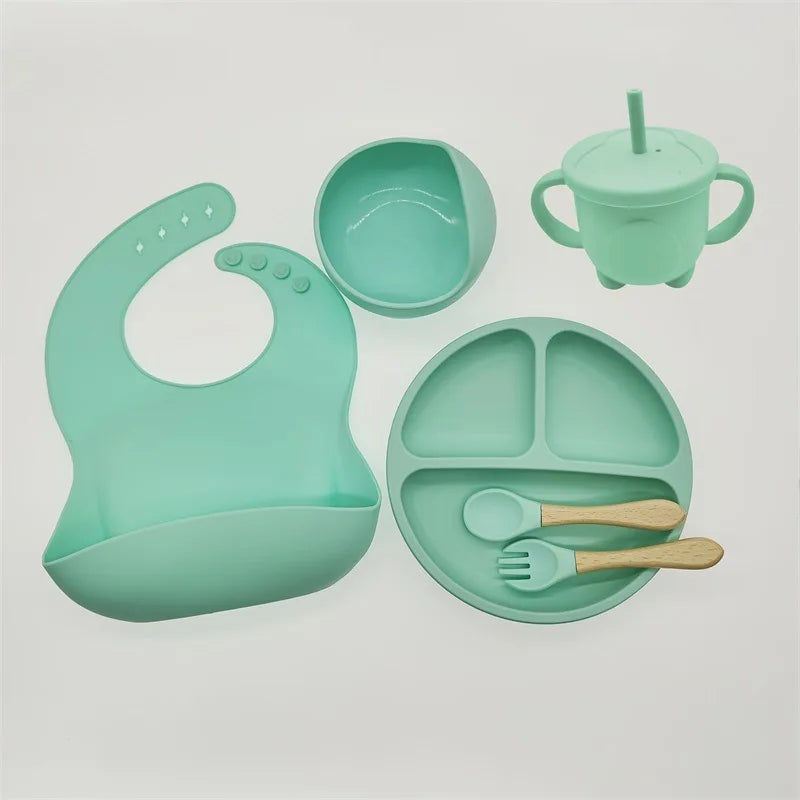 Un ensemble BABY PREMA de couleur sarcelle 6-8 Pièces de Vaisselle pour enfants, fabriqué à partir de silicone de haute qualité, comprend un bavoir, un bol, une tasse avec poignées et paille, une assiette divisée, une cuillère et une fourchette, le tout disposé sur un fond blanc.