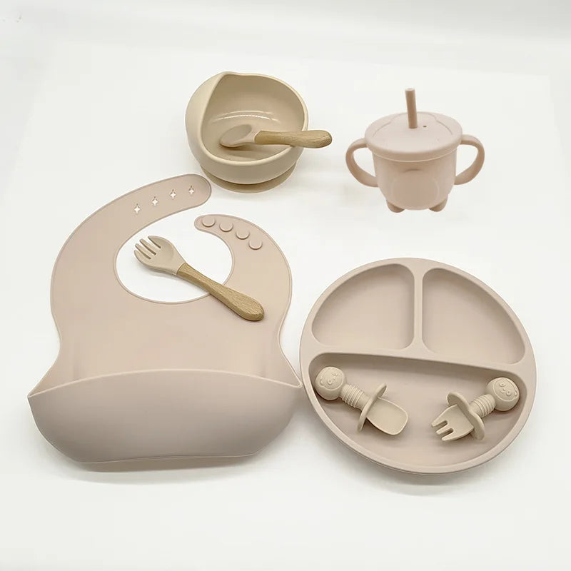 Un ensemble d'alimentation pour bébé comprenant un bavoir, une assiette divisée, un bol, une cuillère, des fourchettes, des tétines et une tasse avec poignées et une paille, le tout en silicone beige clair de haute qualité de l'Ensemble 6-8 Pièces de Vaisselle pour enfants de BABY PREMA.