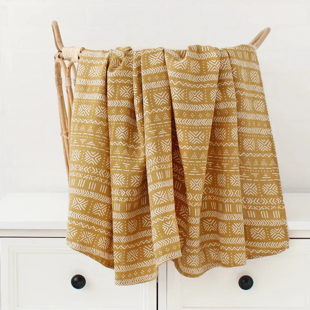 Un pantalon palazzo style bohème jaune moutarde à motifs tribaux blancs et Couvertures en Bambou Bébé | Emmaillotage suspendu sur un cintre en bois sur fond neutre.