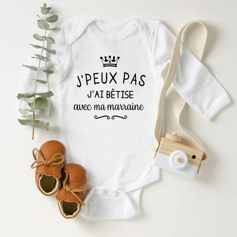 Un Body Blanc Coton Manches Longues de BABY-PREMA avec un graphique de couronne et le texte français "J'PEUX PAS J'AI BÊTISE AVEC MA MARRAINE" repose sur une surface blanche. À côté se trouvent des mocassins marron, un petit appareil photo jouet et un brin de feuillage vert : des vêtements parfaits pour bébés pour votre nouveau-né.