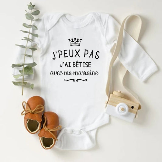 Un Body Blanc Coton Manches Longues de BABY-PREMA avec un graphique de couronne et le texte français "J'PEUX PAS J'AI BÊTISE AVEC MA MARRAINE" repose sur une surface blanche. À côté se trouvent des mocassins marron, un petit appareil photo jouet et un brin de feuillage vert : des vêtements parfaits pour bébés pour votre nouveau-né.