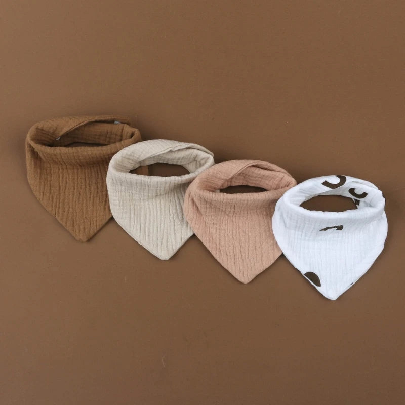 Quatre bavoirs bandana pour bébé sont disposés en rangée sur un fond marron. Ce Bavoirs en mousseline de coton pour bébé de BABY-PREMA se décline dans les couleurs marron, beige, rose et blanc avec un motif de taches brunes. Fabriqués à partir de bavoirs en mousseline de coton, les bavoirs ont un aspect tissu texturé et sont soigneusement pliés.