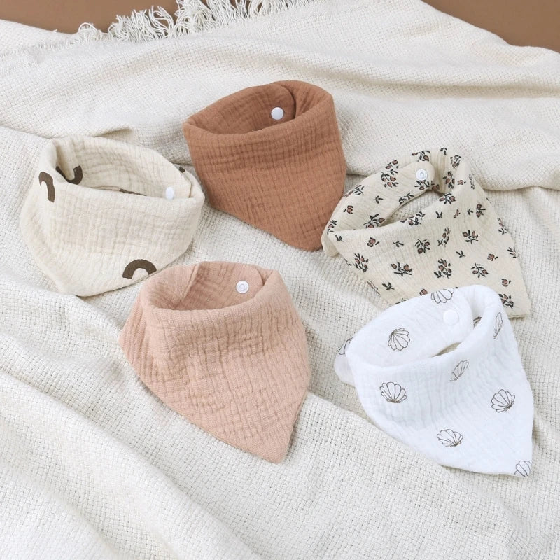 Cinq doux Bavoirs en mousseline de coton pour bébé BABY-PREMA disposés en demi-cercle sur un tissu texturé. Les bavoirs sont de différents motifs et couleurs : beige uni, marron uni, blanc à imprimés fleuris, beige à imprimés fleuris et blanc à petits motifs marron. Fabriqué en mousseline de coton pour un confort ultime.