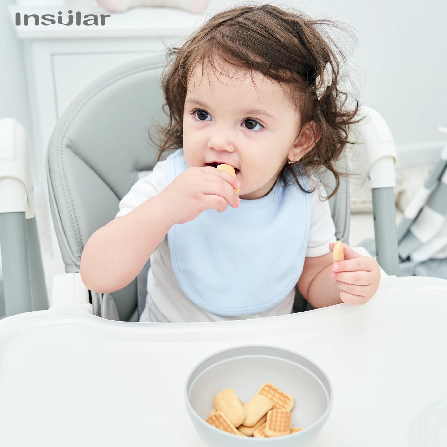 Un tout-petit profite du goûter dans une chaise haute avec un bol de biscuits et le Bavoir Bébé en Coton Bio BABY PREMA.