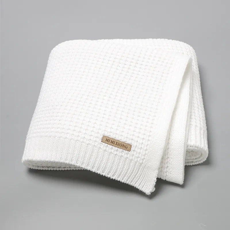 Une couverture en tricot blanc pliée, parfaite comme Couverture Bébé Tricot Fin 90*70cm - Baby-Prema, est présentée sur un fond uni gris. Il comporte une petite étiquette beige intitulée « Baby-Prema.