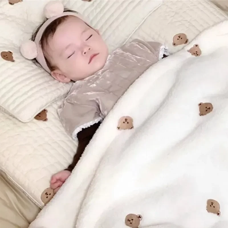 Un bébé paisible dort paisiblement, orné d'un joli bandeau, enveloppé dans une couverture BABY PREMA Couverture de Bébé en Molleton de Corail avec d'adorables détails de petit ours, entouré de nécessaire pour bébé.