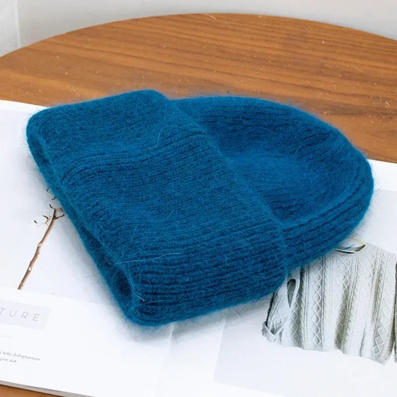 Un bonnet Angora Pour Maman en laine bleue et douillette de BABY PREMA posé sur un magazine ouvert avec des textures de bois, dégageant une ambiance chaleureuse et tendance comme accessoire bébé parfait.