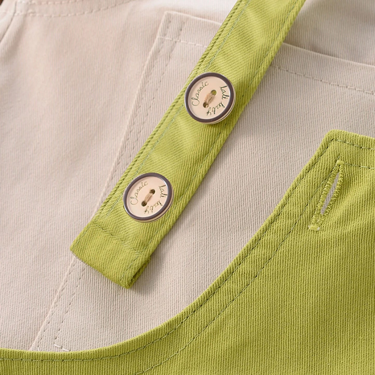 Gros plan sur une salopette vert clair (Salopette Bébé Unie) avec détails de boutons. Les boutons sont ronds, métalliques et gravés des mots « Classic » et « Ready ». Cette salopette pour enfants de BABY-PREMA possède une poche et une doublure beige, confectionnée en coton doux.