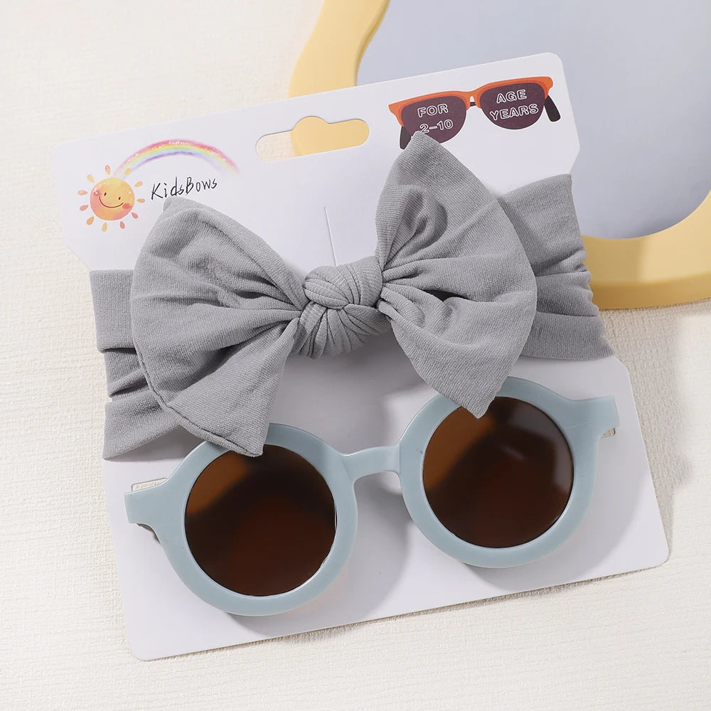 Un ensemble d'accessoires pour enfants exposé, comprenant un serre-tête gris avec un nœud et une paire de lunettes de soleil rondes grises. L'emballage indique « BABY PREMA - Set d'Accessoires Bébé | Lunettes de Cheveux pour Bébé » et indique qu'il s'agit d'accessoires pour cheveux de bébé âgés de 1 à 6 ans.