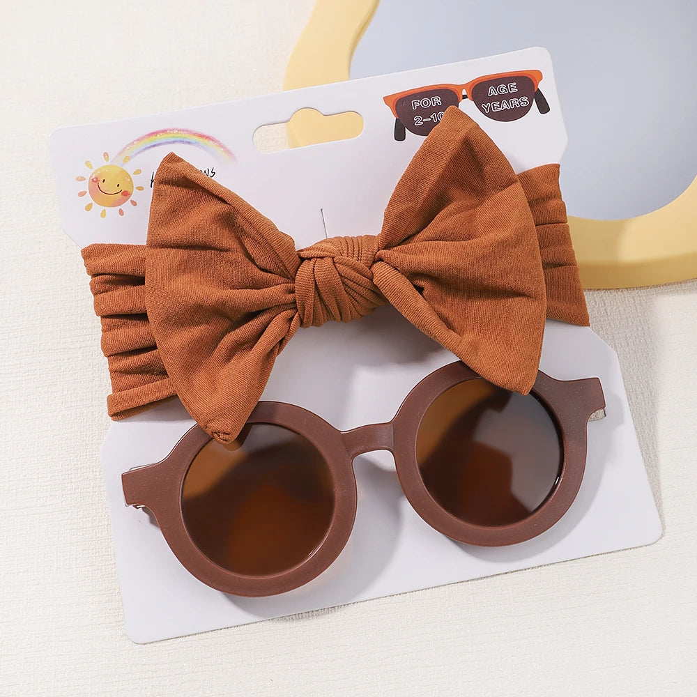 Un ensemble d'accessoires pour enfants, dont un Set d'Accessoires Bébé rond marron | Lunettes de Cheveux pour Bébé BABY PREMA et bandeaux à nœuds en nylon assortis avec un grand nœud, présentés sur un emballage blanc.