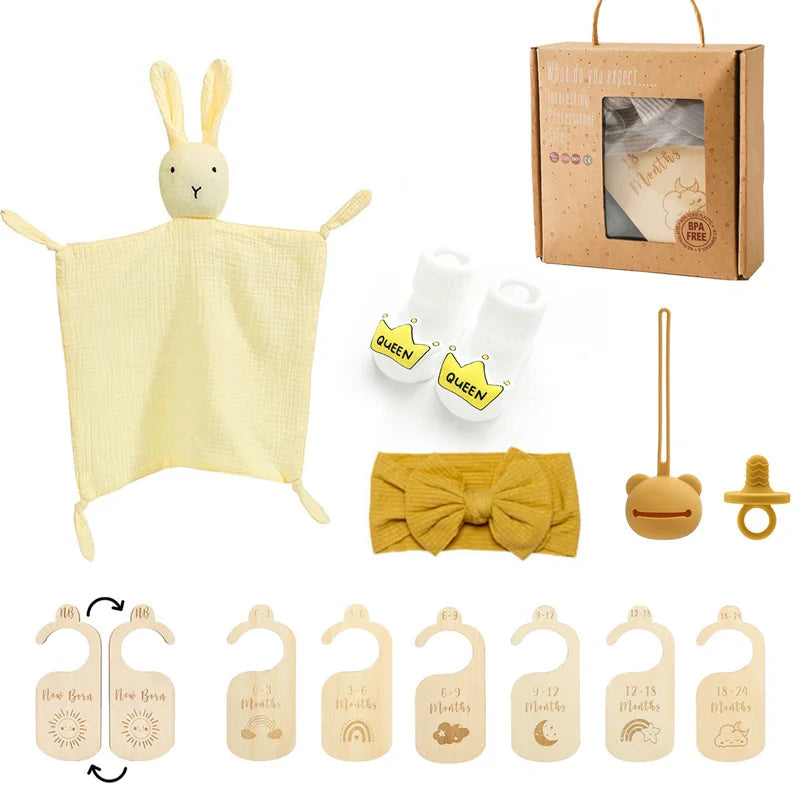 Une collection d'essentiels pour l'hygiène de bébé et d'accessoires dont un doudou BABY PREMA Coffret Cadeau Naissance Bébé, des cartes jalons en bois, une paire de chaussettes avec écrit "reine" sur les semelles, un bandeau jaune.