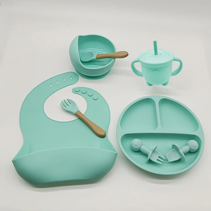 Un ensemble d'articles d'alimentation pour bébé vert menthe en silicone de haute qualité, comprenant un bavoir, un bol avec une cuillère, une tasse avec anses et paille, une assiette divisée, une fourchette et deux tétines de l'Ensemble BABY PREMA 6-8 La collection Pièces de Vaisselle pour enfants est disposée sur une surface blanche.