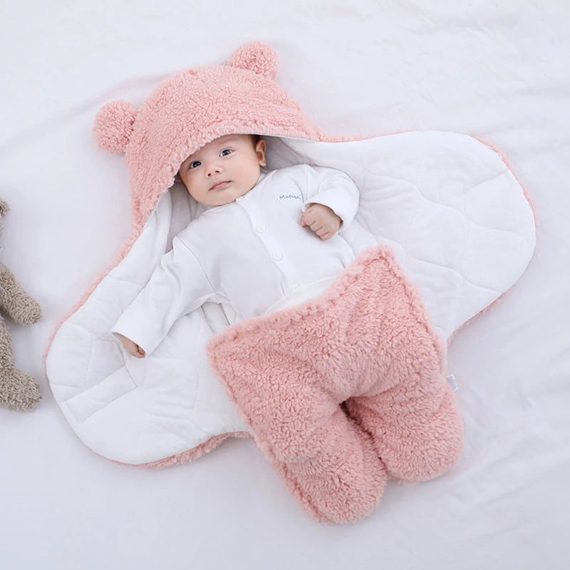 Un bébé douillet enveloppé dans la Couverture bébé Nid D'Ange de BABY-PREMA, complété par un joli chapeau à oreilles d'ours et de petits chaussons assortis.