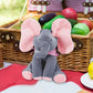 Un éléphant en peluche BABY-PREMA avec de grandes oreilles roses assis devant un panier de pique-nique rempli d'œufs colorés sur une couverture de pique-nique légère à l'extérieur.