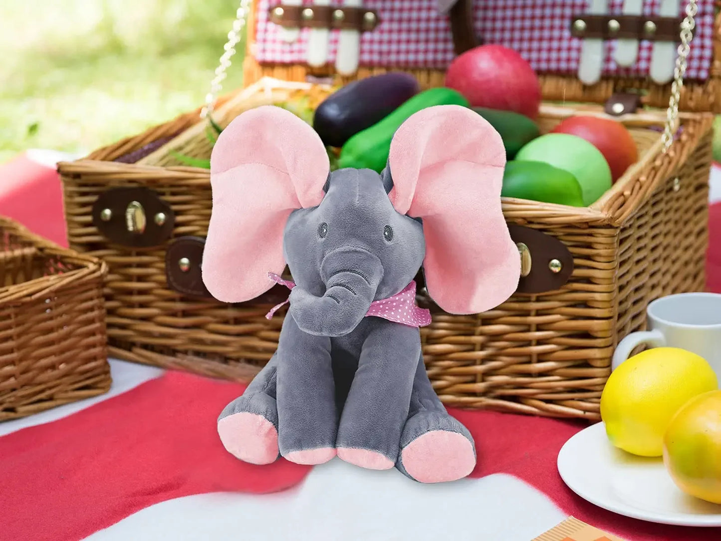 Un éléphant en peluche BABY-PREMA avec de grandes oreilles roses assis devant un panier de pique-nique rempli d'œufs colorés sur une couverture de pique-nique légère à l'extérieur.