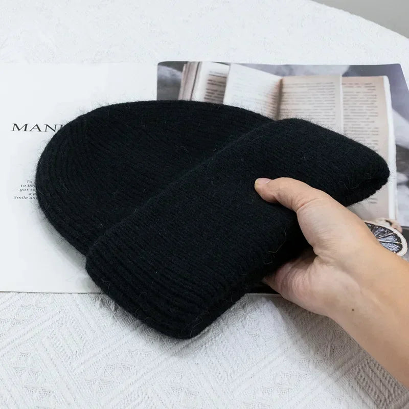 Une main tenant un bonnet pour bébé Bonnet Angora Pour Maman noir plié au-dessus d'un magazine ouvert, placé sur une surface blanche texturée par BABY PREMA.