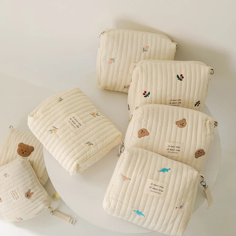 Une collection de jolies pochettes BABY PREMA Sac à Couches en Coton pour poussette aux couleurs pastel, chacune ornée de motifs de broderie simples mais adorables tels que des fleurs, des ours et du texte minimaliste, avec de délicats accents de plumes.