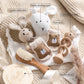 Un arrangement douillet de Coffret Cadeau Naissance Bébé et de jouets, dont un hochet lapin au crochet, une serviette baveuse, des chaussettes de sol, des jouets en bois et un jalon, le tout joliment disposé sur un tapis moelleux. Marque : BABY PREMA