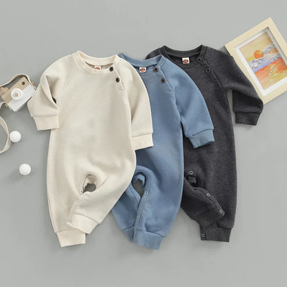 Trois combinaisons à manches longues pour bébé beige, bleu et gris foncé sont disposées sur une surface grise. Ces Genouillères Barboteuses | Naissance Bébé de BABY PREMA est en tissu gaufré doux. A proximité se trouvent deux balles de ping-pong, un appareil photo jouet et un cadre photo avec une peinture de paysage.