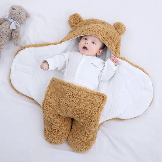Un bébé a l'air surpris et adorable, bien enveloppé dans une couverture BABY PREMA Cocoon Nid d'Ange bébé avec de petites oreilles, des chaussons en pattes d'ours, sur un fond blanc, avec une peluche à proximité.