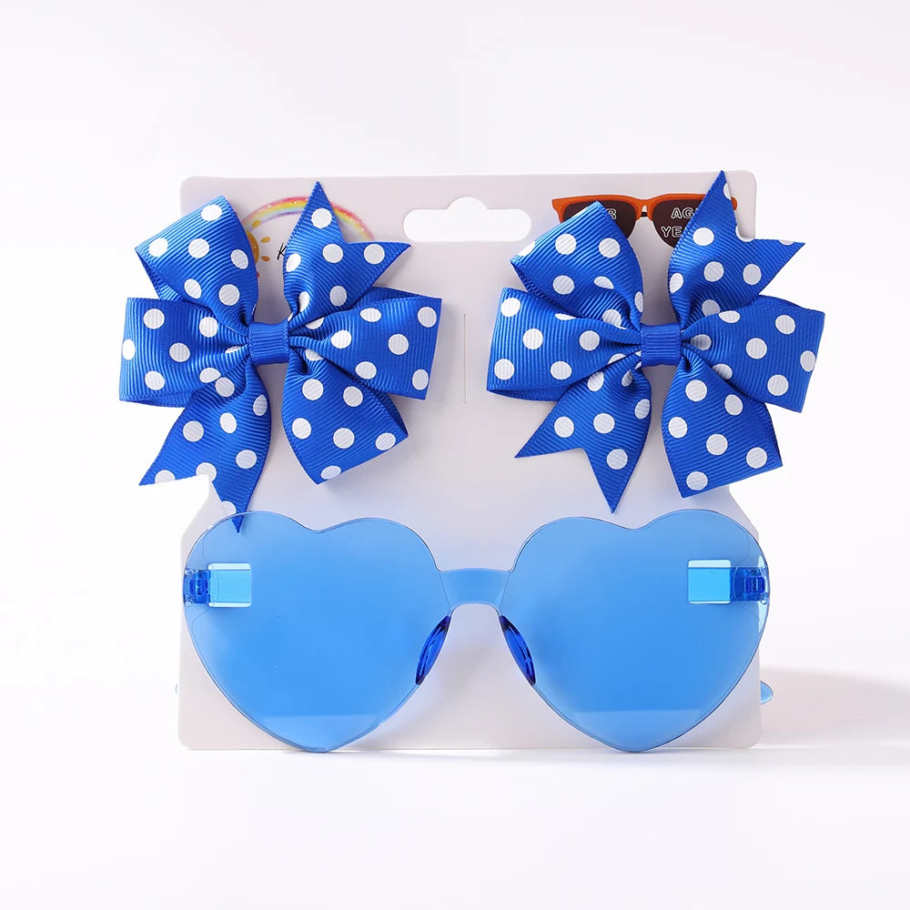 Une paire de lunettes de soleil pour bébé bleues en forme de cœur et deux nœuds pour cheveux bleus à pois blancs, attachés à une carte présentoir blanche du Set d'Accessoires Bébé | Collection Lunettes de Cheveux pour Bébé de BABY PREMA.