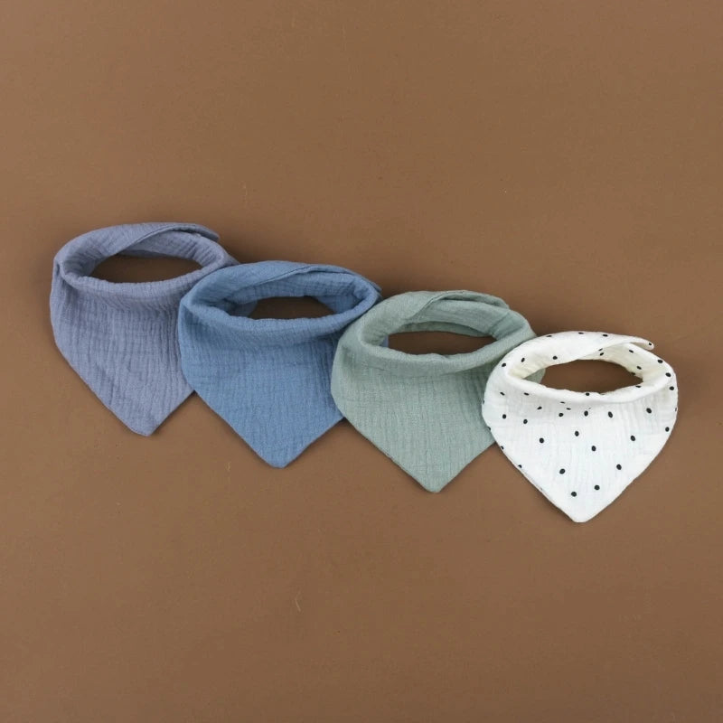 Un lot de 4 Bavoirs en mousseline coton pour bébé de BABY-PREMA est disposé en rangée sur un fond marron. Les bavoirs en mousseline de coton sont de gauche à droite : bleu clair, bleu foncé, vert sauge et blanc à pois noirs. Chaque bavoir est triangulaire avec une encolure arrondie.