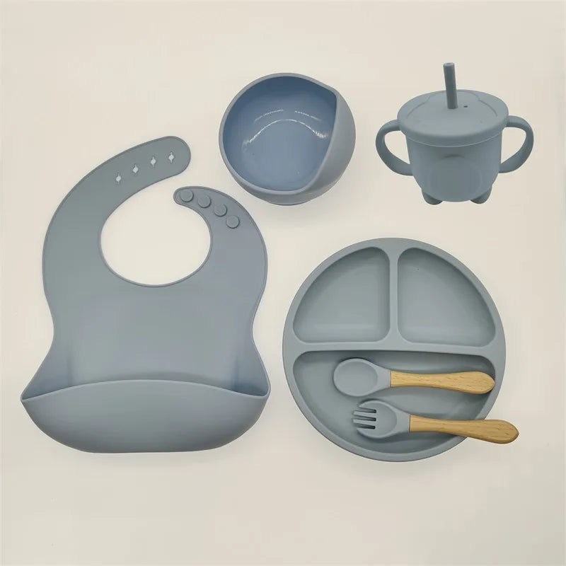 Un ensemble d'articles d'alimentation pour bébé de couleur bleu tendre, comprenant un bavoir, un bol, un gobelet, une assiette divisée, une cuillère et une fourchette avec des manches en bois en silicone de haute qualité, disposés sur un fond blanc. The BABY PREMA Ensemble 6-8 Pièces de Vaisselle pour enfants.