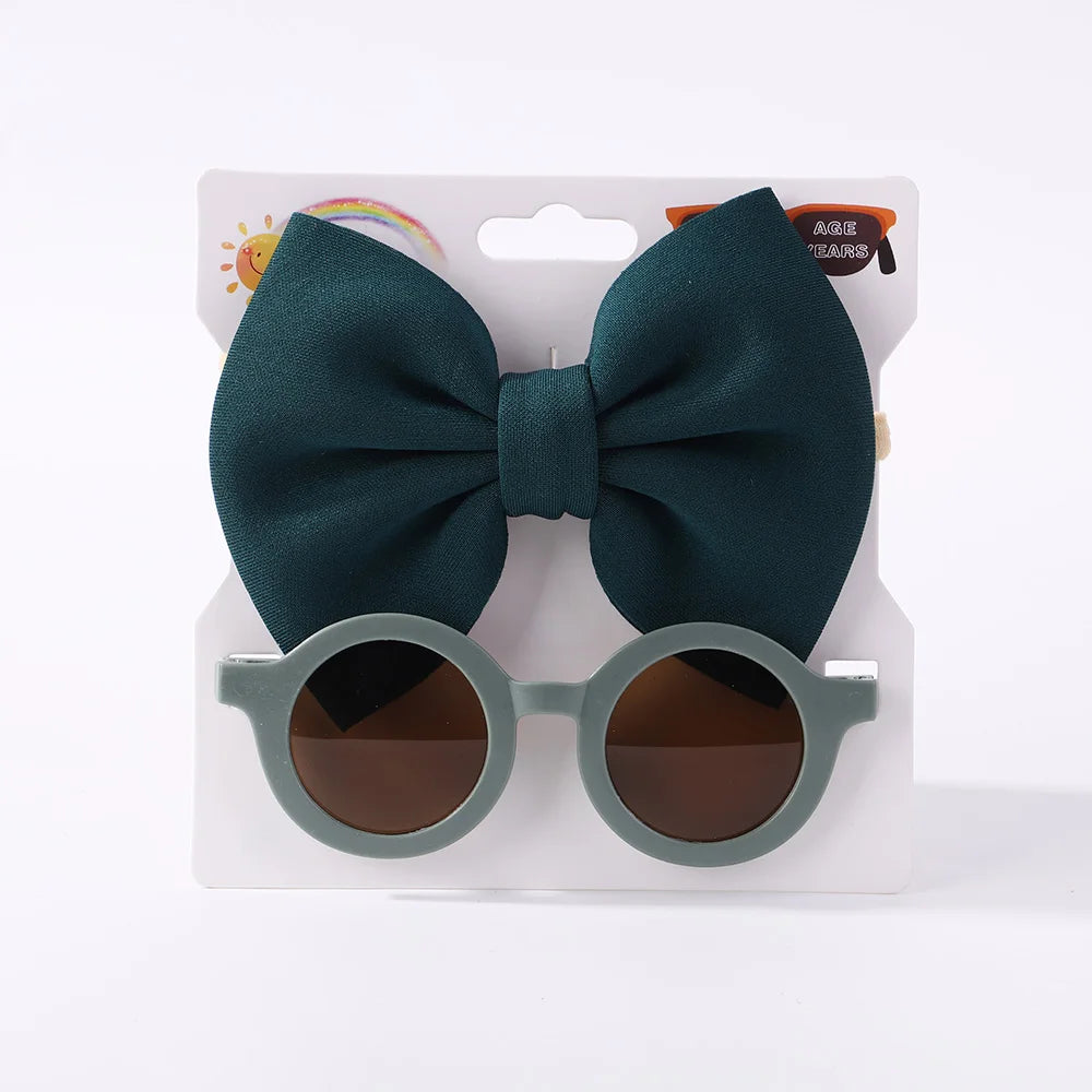 Une paire de lunettes de soleil rondes avec une monture vert grisâtre est présentée avec un grand nœud papillon vert attaché en haut, parfait pour ajouter du style à l'ensemble de votre bébé. Complétez le look avec l'Ensemble d'Accessoires Bébé | Lunettes de Cheveux pour Bébé de BABY PREMA pour un style charmant et coordonné.