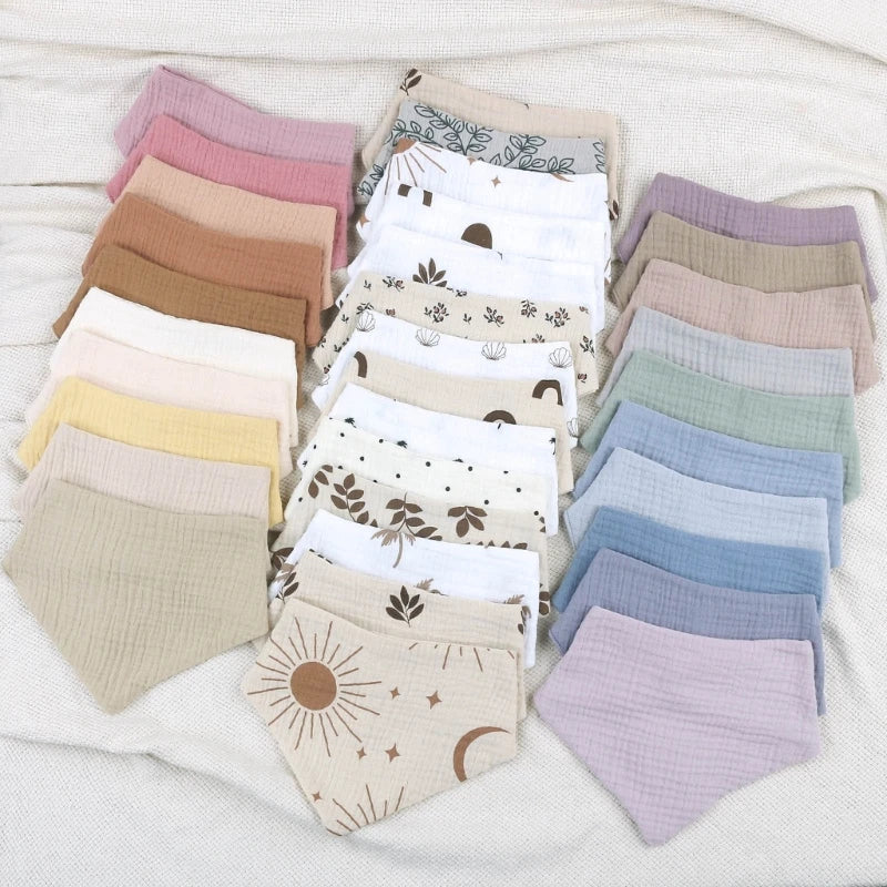 Une collection de bavoirs pliés soigneusement disposés sur une surface blanche. Les Bavoirs en mousseline coton pour bébé de BABY-PREMA présentent différentes couleurs, dont des nuances de rose, marron, jaune, blanc, bleu et violet. Certains bavoirs ont des motifs comme des fleurs, du soleil, de la lune et des étoiles, tandis que d'autres sont de couleur unie. Fabriqué en mousseline de coton douce pour des vêtements de protection doux.