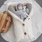 Un paquet de joie douillet : un bébé content, confortablement enveloppé dans une couverture tricotée de couleur crème avec des accessoires pour bébé, complété par un élégant sac en cuir marron à côté. Couverture de Poussette Pour Bébé de BABY PREMA.