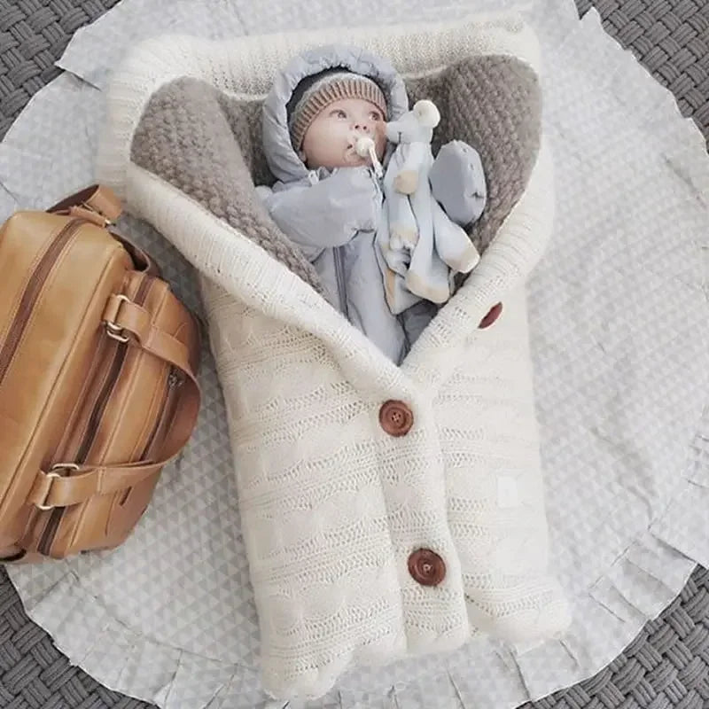 Un paquet de joie douillet : un bébé content, confortablement enveloppé dans une couverture tricotée de couleur crème avec des accessoires pour bébé, complété par un élégant sac en cuir marron à côté. Couverture de Poussette Pour Bébé de BABY PREMA.