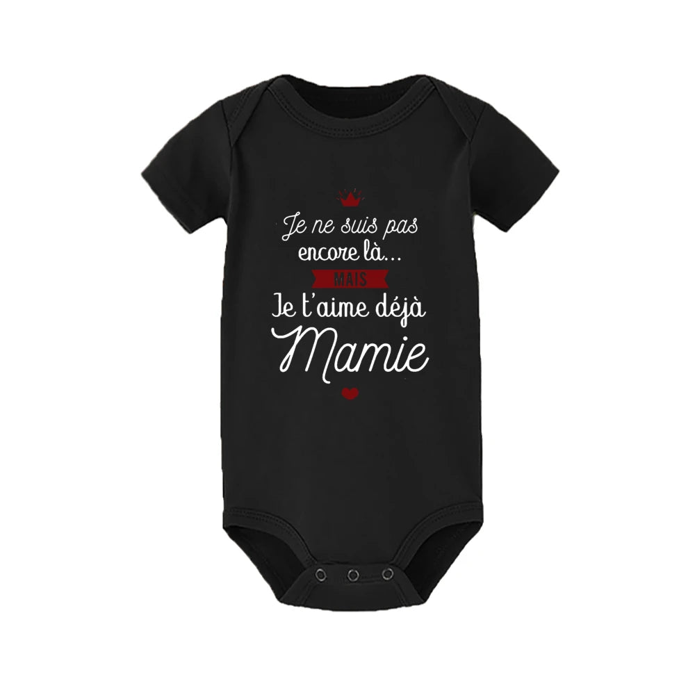 Un Body Coton été Imprimé noir par BABY-PREMA, parfait comme body d'été, avec le texte en français qui dit "Je ne suis pas encore là... MAIS Je t'aime déjà Mamie" avec des éléments décoratifs comme un petit rouge coeur et un diadème rouge. La combinaison a des manches courtes et des boutons-pression en bas, idéale pour votre bébé.