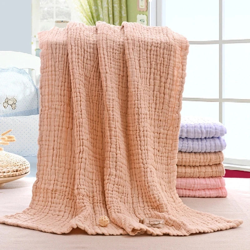 Une couverture texturée douce et moelleuse de couleur pêche élégamment drapée sur une surface, avec des piles de BABY PREMA Couvertures Mousseline | 6 Couches pour Nouveau-né dans des tons pastel de bleu, rose et violet en fond.