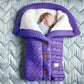 Un nouveau-né qui dort paisiblement enveloppé dans une douillette gigoteuse BABY PREMA Couverture de Poussette Pour Bébé en tricot violet et blanc en forme de veste avec des boutons en bois.