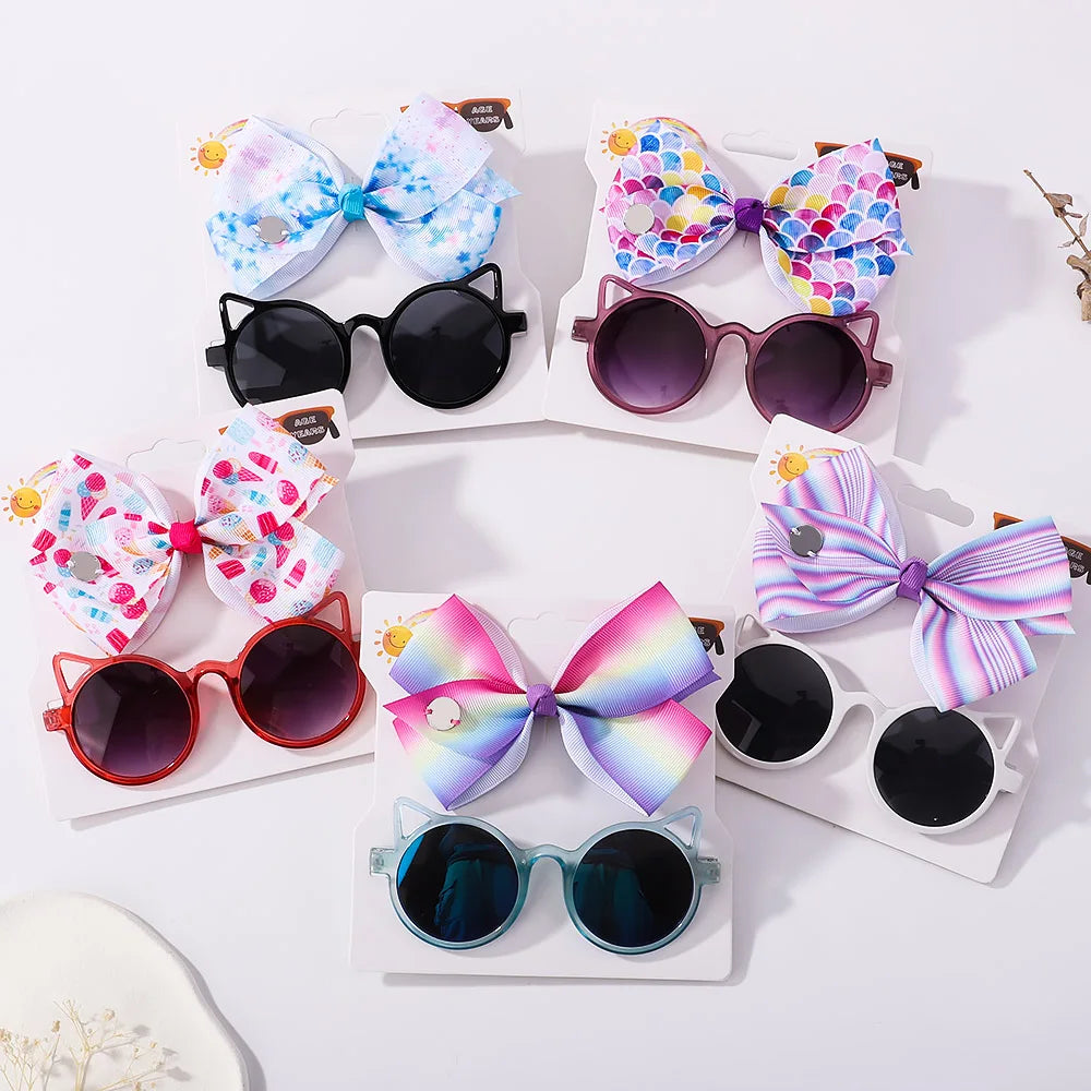 Présentoir de six paires de Set d'Accessoires Bébé coloré | Lunettes de Cheveux pour Bébé de BABY PREMA et bandeaux à nœuds en nylon assortis, disposés à plat. Chaque paire comprend différentes couleurs de monture et des motifs fantaisistes sur les nœuds.