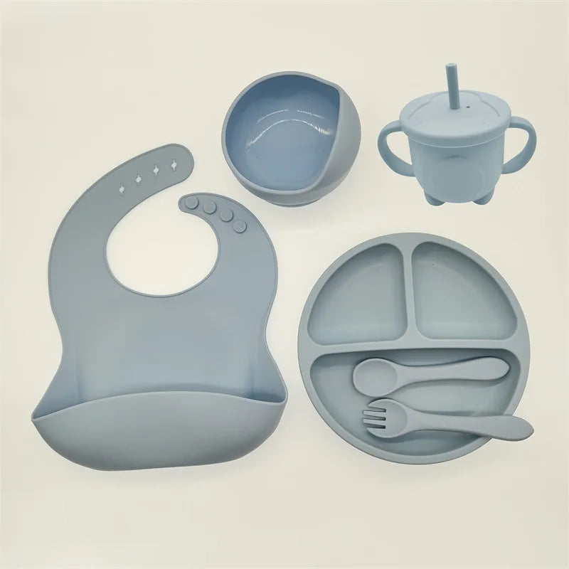 Un ensemble BABY PREMA 6-8 Pièces de Vaisselle pour enfants en bleu clair, comprenant un bavoir, un bol, un gobelet avec poignées et paille, une assiette divisée, une cuillère et une fourchette, le tout fabriqué en silicone de haute qualité pour plus de durabilité et de sécurité.