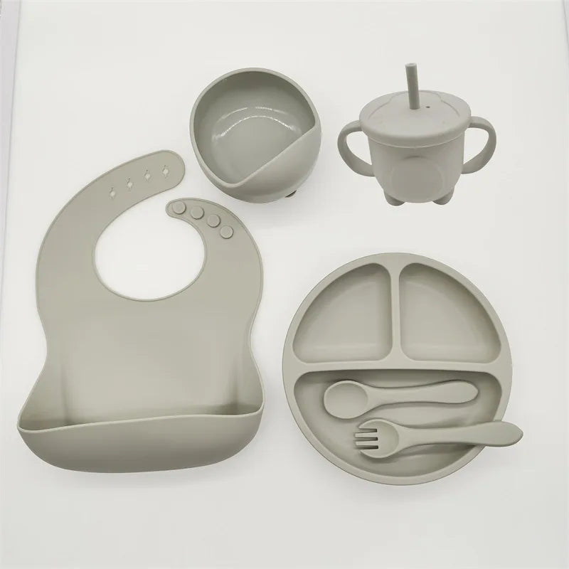 Un ensemble d'articles d'alimentation pour bébé en silicone gris de haute qualité, comprenant un bavoir, des bols à ventouse, un gobelet, une assiette divisée, une fourchette et une cuillère disposés sur un fond blanc. Le produit est l'Ensemble 6-8 Pièces de Vaisselle pour enfants de BABY PREMA.