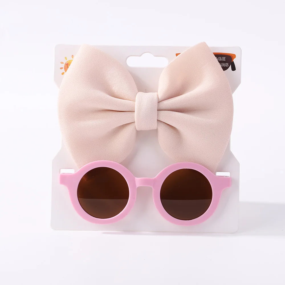 Un ensemble d'accessoires bébé | Lunettes de Cheveux pour Bébé de BABY PREMA avec verres foncés attachés à un emballage comportant un grand nœud beige clair.