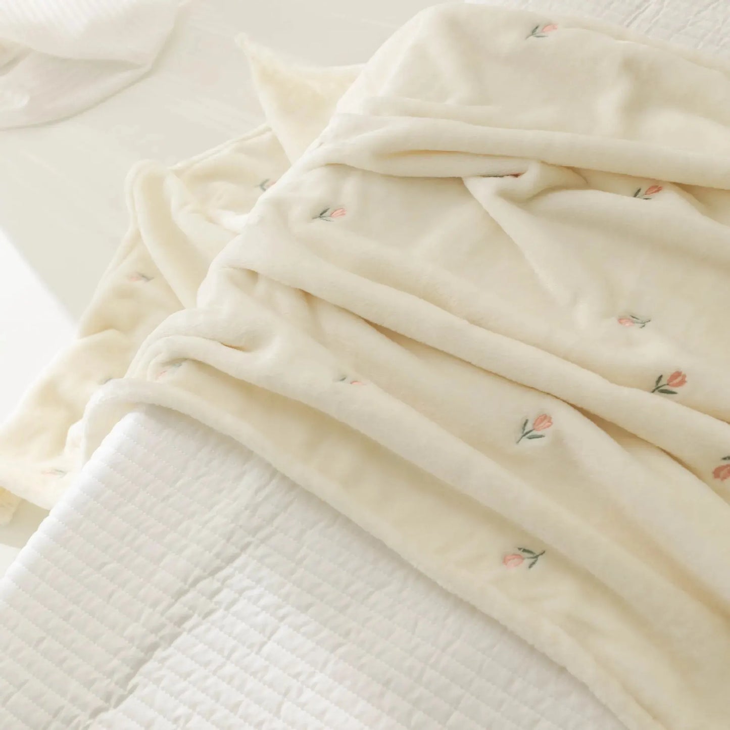 Couverture de Bébé en Molleton de Corail BABY PREMA douce et confortable avec une délicate broderie florale, délicatement pliée sur un couvre-lit blanc texturé, invitant une sensation de chaleur et de tranquillité dans la chambre. Parfait dans le cadre des nécessaires pour