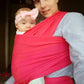 Une femme au doux sourire porte un bébé dans un BABY-PREMA Porte-Bébé rouge vif dès la Naissance | Écharpe de Portage. Le bébé, portant un bandeau rose clair avec un nœud, a l'air bien.