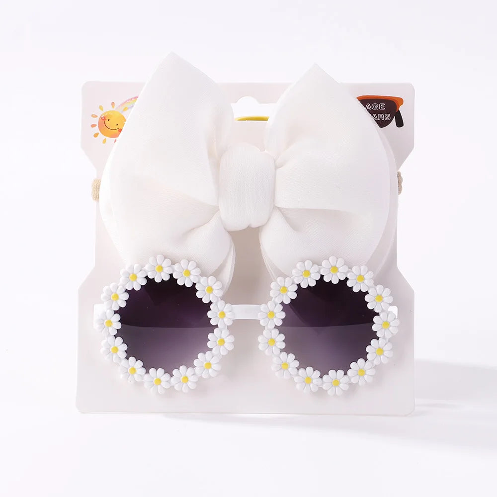 Des lunettes de soleil rondes pour bébé, décorées d'un motif de marguerite autour des verres, sont présentées sur un support en carton blanc avec un grand nœud blanc fixé au-dessus de l'ensemble d'accessoires bébé | Lunettes de Cheveux pour Bébé de BABY PREMA.