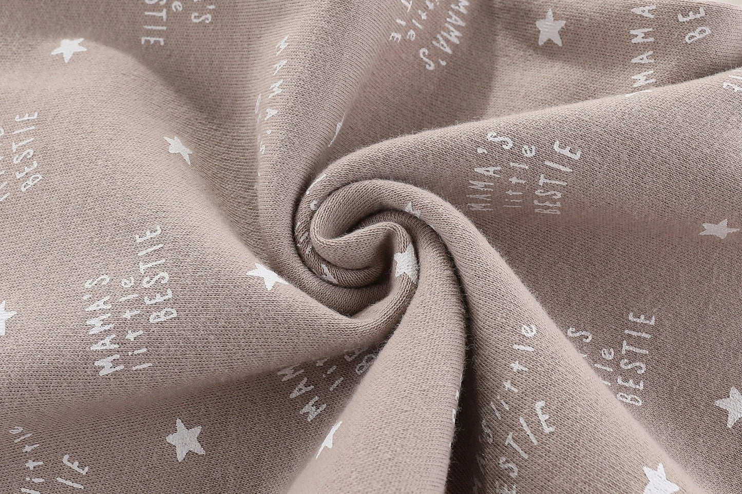 Gros plan sur un tissu beige avec un motif d'étoiles et le texte répété "Mama's little bestie" en blanc. Le tissu est froncé au centre, créant un tourbillon. Un choix idéal pour bébé, ce Lot 6 Body Coton Bébé Manches Courtes de BABY PREMA constitue un cadeau de naissance parfait.