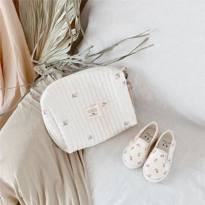 Une pose plate élégante comprenant un Sac de Couches Rangement Bébé de BABY PREMA avec une délicate broderie florale à côté d'une adorable paire de petites chaussures de bébé avec un motif floral assorti, le tout disposé sur un fond doux et aux tons neutres.