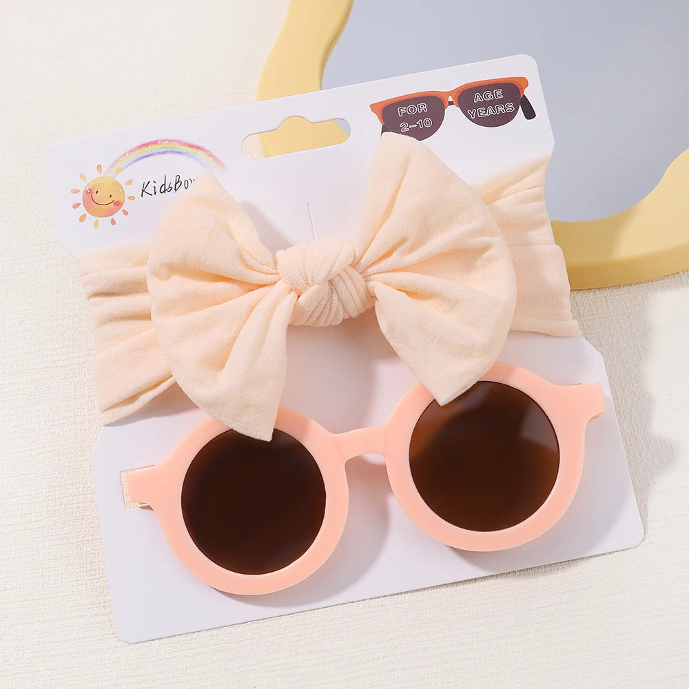 Une paire de lunettes de soleil pour bébé rondes roses avec des verres foncés et un bandeau crème avec un nœud sont emballés ensemble. L'emballage est étiqueté pour les enfants de 0 à 3 ans, des accessoires parfaits pour cheveux de bébé. Cet adorable ensemble s'appelle Set d'Accessoires Bébé | Lunettes de Cheveux pour Bébé de BABY PREMA.