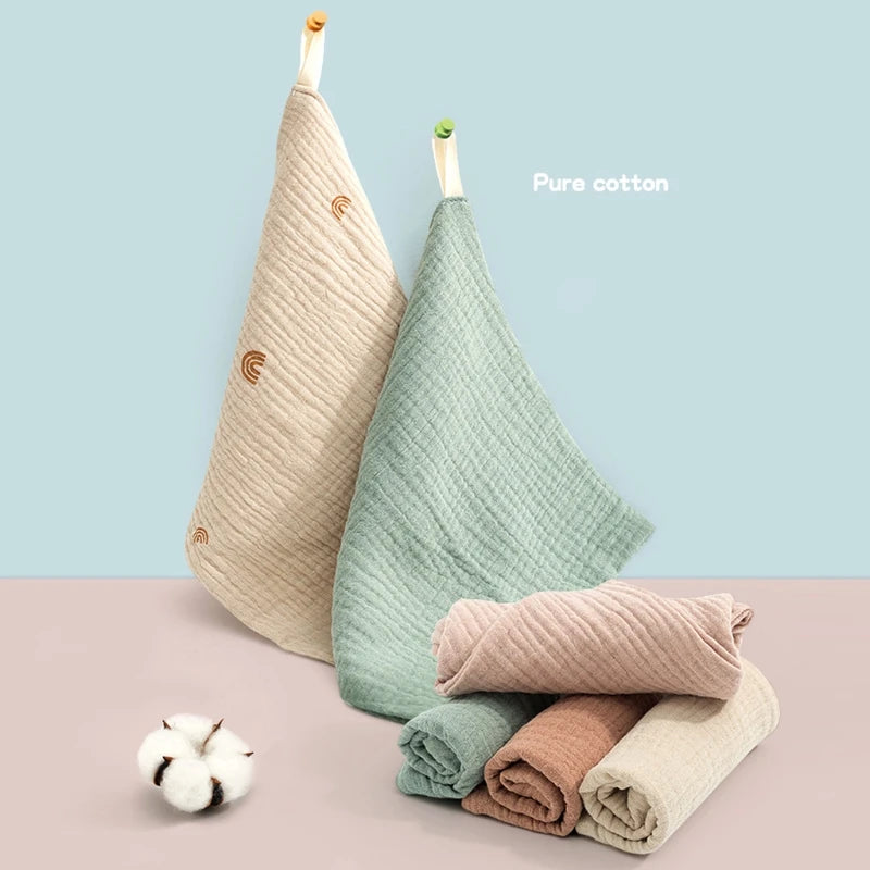 Deux serviettes suspendues et un ensemble empilé de serviettes en coton léger BABY PREMA aux couleurs douces et pastel avec un cotonnier à côté, sur un fond bicolore, mettant en valeur la matière naturelle et pur coton idéale pour le Lot de 5 Langes Bébé en Coton.