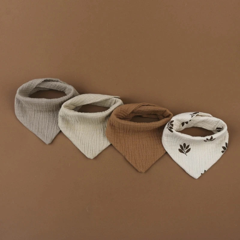 Quatre bavoirs bandana bébé sont exposés sur fond marron. Ces Bavoirs en mousseline coton pour bébé de BABY-PREMA comprennent des bavoirs disposés côte à côte dans des couleurs et des motifs variés : gris clair, crème, marron et blanc avec des imprimés de feuilles marron. Ces bavoirs en mousseline de coton offrent à la fois style et confort à votre tout-petit.