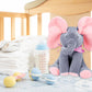 Un jouet éléphant en peluche doux BABY-PREMA est posé sur la table à langer d'un bébé prématuré, entouré d'une paire de chaussures pour bébé, d'un biberon et de hochets, créant un cadre de chambre de bébé confortable et nourrissant.